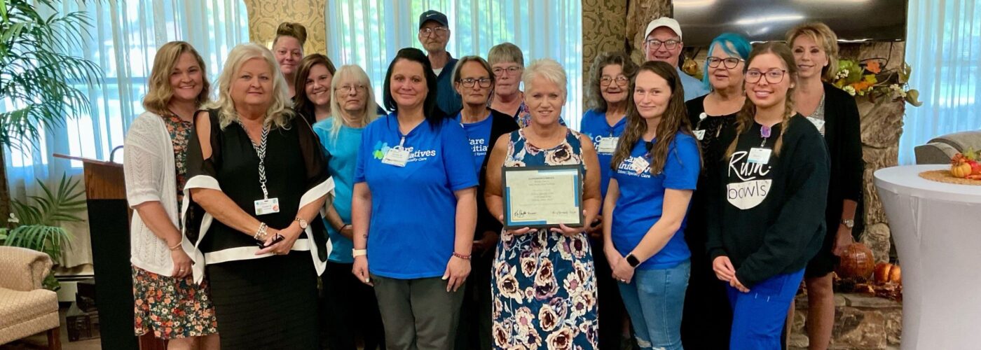 A group of people stands in a room with large windows and plants. A woman in the center holds a certificate, surrounded by casually dressed individuals, some wearing matching blue shirts. They stand before a stone wall with a TV screen and podium, celebrating their Care Initiative.