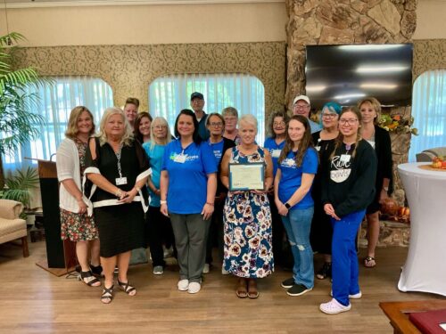 A group of people stands in a room with large windows and plants. A woman in the center holds a certificate, surrounded by casually dressed individuals, some wearing matching blue shirts. They stand before a stone wall with a TV screen and podium, celebrating their Care Initiative.