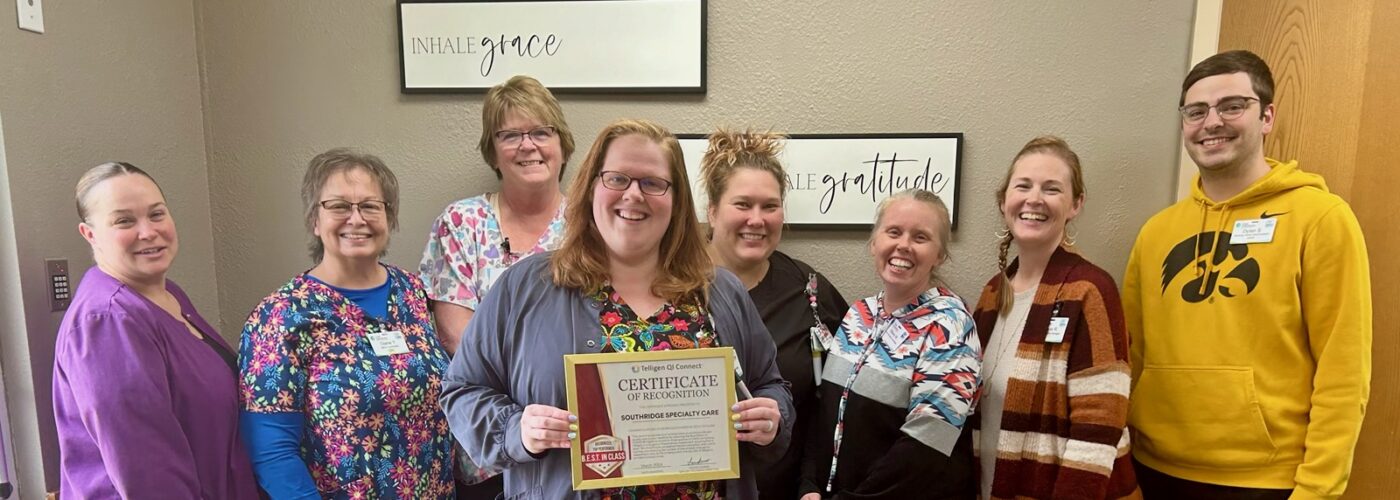 A group of eight people stands indoors, smiling warmly. One person proudly holds a certificate, showcasing their participation in the Care Initiative. They are dressed in casual and colorful work attire against light walls with framed motivational signs.