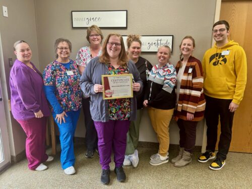 A group of eight people stands indoors, smiling warmly. One person proudly holds a certificate, showcasing their participation in the Care Initiative. They are dressed in casual and colorful work attire against light walls with framed motivational signs.