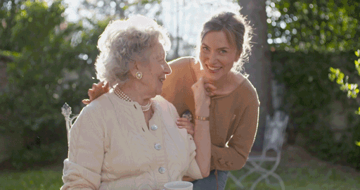 A smiling younger woman embraces an older woman seated outdoors, both enjoying a sunny day in the garden. With care and warmth, they share a joyful moment as the older woman holds a cup. Brought to you by Care Initiative.