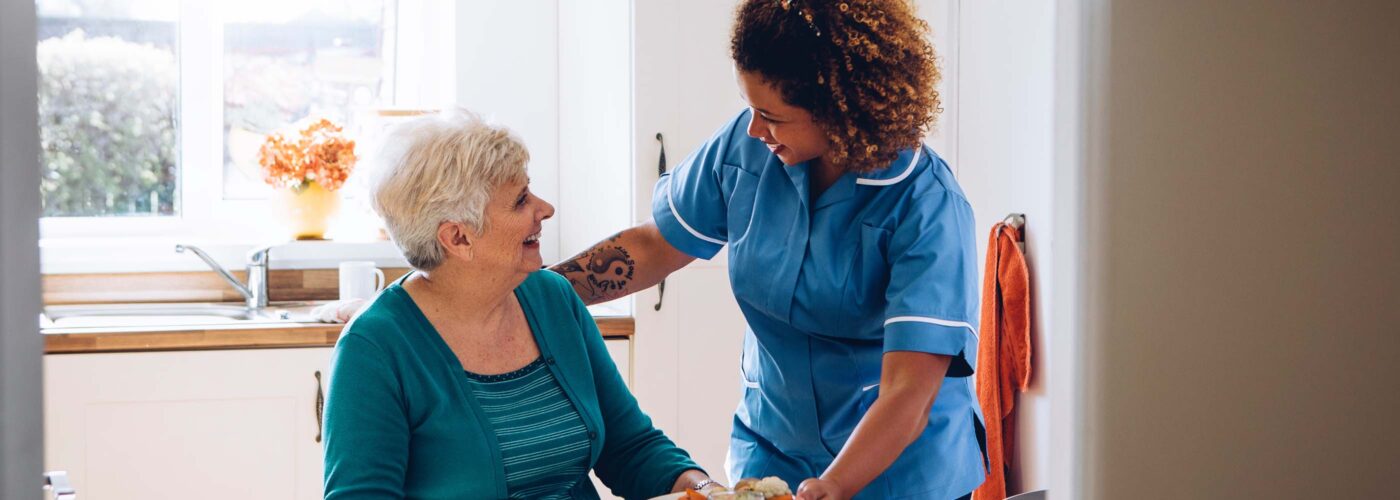 A caregiver in a blue uniform serves a meal to an elderly woman at a kitchen table, embodying the Care Initiative. The bright room features a window in the background, and their smiles create a warm and friendly atmosphere.