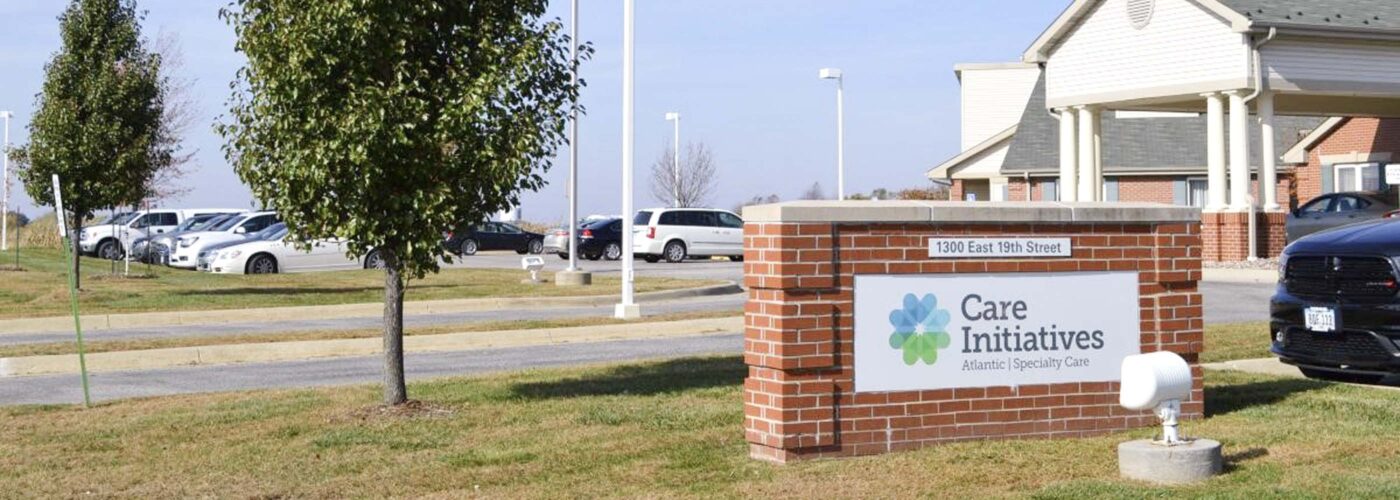 The exterior view features a building with a brick sign reading Care Initiative. The parking lot is partially visible with several cars, and an American flag flies on a pole. Green lawns and trees provide a backdrop under the clear sky. The brand name, Care Initiative, is prominently displayed on the sign.