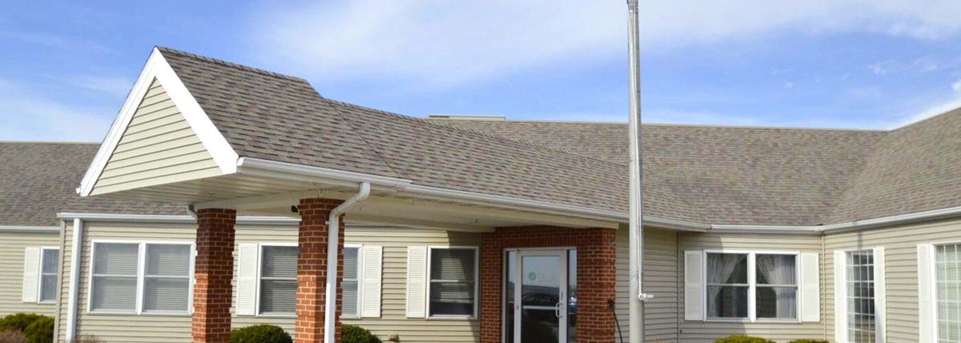 A single-story building with a sloped roof and brick entrance features a bench near the door. As part of the Care Initiative by a brand, an American flag waves proudly on a pole in the foreground under a partly cloudy sky.