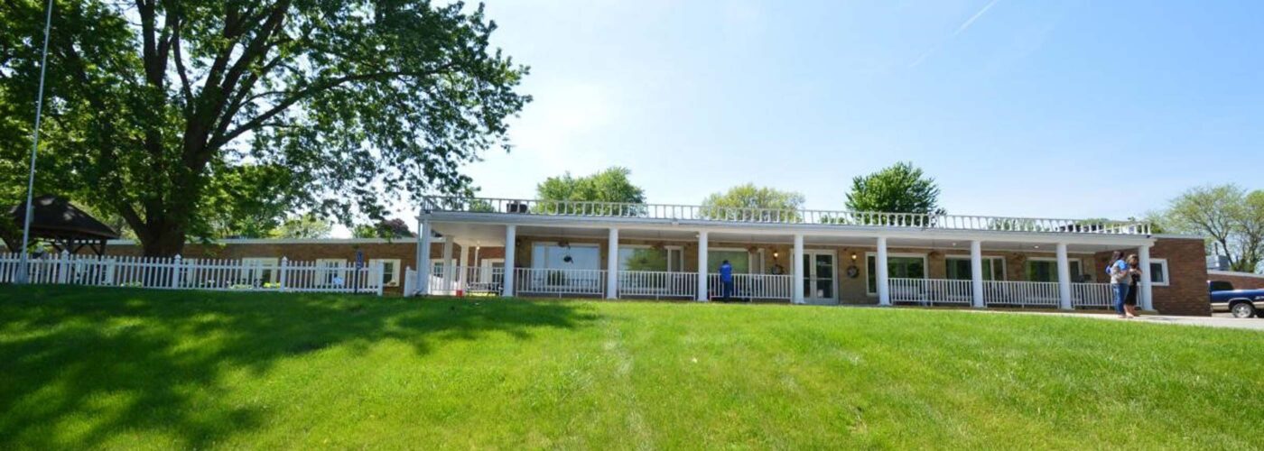 A single-story white building with a porch and white railings sits atop a grassy hill on a sunny day. Several people stroll nearby, while large trees provide shade in the background, embodying the spirit of Care Initiative.