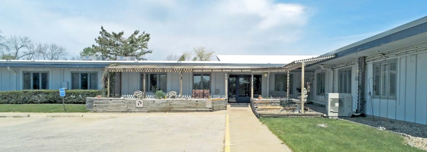 The Care Initiative center is a single-story building with a flat roof, surrounded by a parking area and green lawn. The entrance features a covered walkway and large windows, framed by shrubs and trees in the background under a blue sky with clouds.