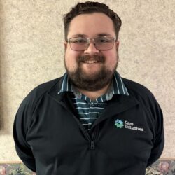 A person with glasses, a beard, and short hair is smiling in a black jacket featuring a Care Initiative logo over a blue and white striped shirt. The background is a beige wall with a floral border.