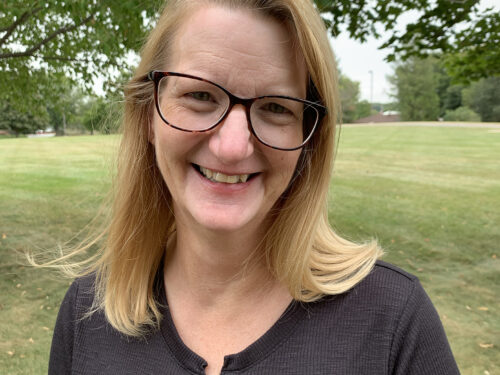 A woman with long blonde hair and glasses is smiling in an outdoor setting. She wears a black top with a lace-up neckline. Green grass and trees are in the background. Brand name: Care Initiative.