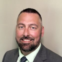 A bearded man with short hair is smiling at the camera. He is wearing a gray suit jacket, a light green shirt, and a dark patterned tie. The background is a plain light-colored wall. Care Initiatives.