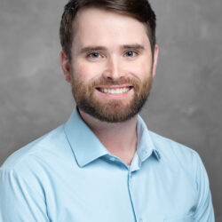 A man with a beard and short brown hair is smiling warmly, dressed in a light blue shirt against a gray background, embodying the spirit of Care Initiative.