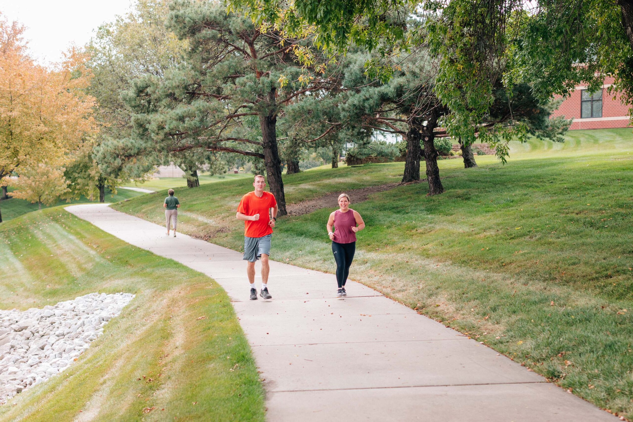 A man and woman are jogging on a paved path through a park, surrounded by green grass and trees. As part of the local Care Initiative, they enjoy the benefits of staying active. Another person is in the distance. The clear sky suggests a pleasant day.