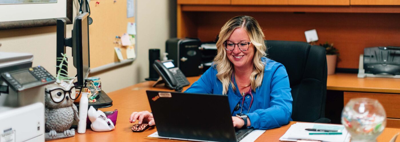 A woman with glasses and blonde hair works at her desk in an office, focusing on a project for Care Initiative. She uses a laptop and smiles warmly. The office is adorned with a painting, a bulletin board, and various desk items, including an owl ornament and a phone.