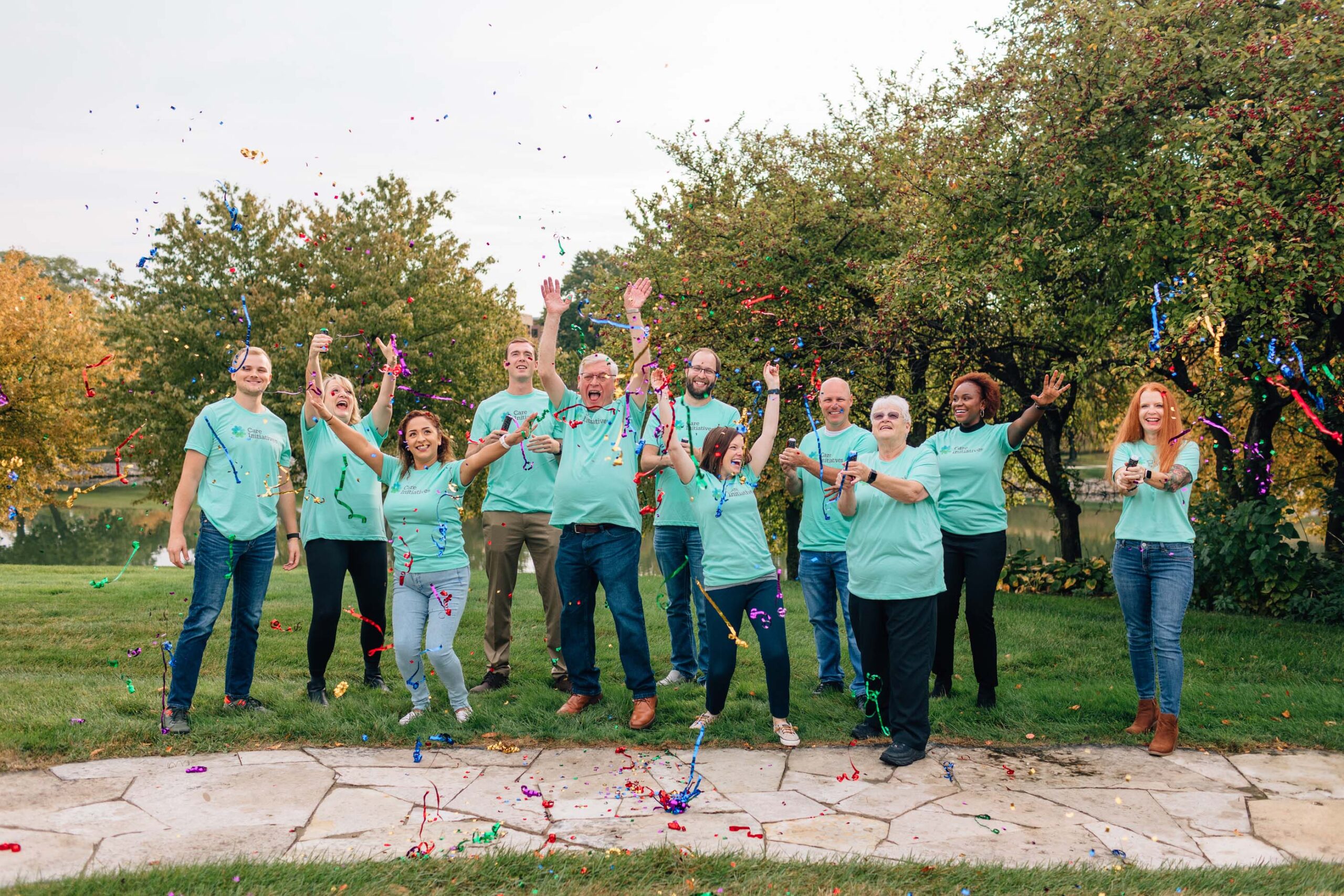 A group of people wearing matching teal shirts stands outdoors on a grassy area, celebrating Care Initiative. They smile with arms raised as colorful streamers fly in the air, with trees and a path visible in the background.