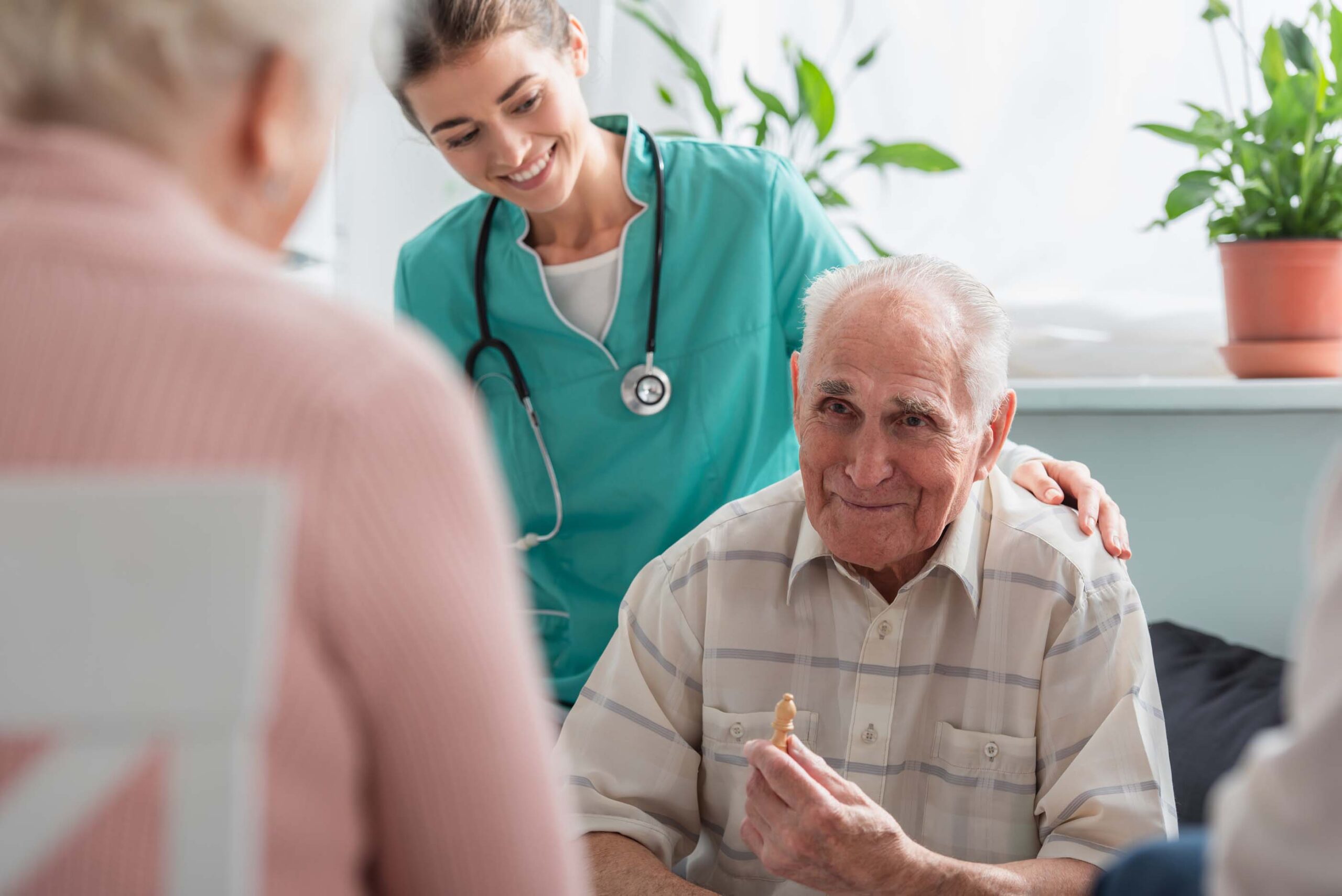 A smiling healthcare worker in teal scrubs with a stethoscope gently rests a hand on the shoulder of an elderly man holding a piece of food. This scene captures the essence of Care Initiative. A woman is with them, partly visible from behind, while plants decorate the background.