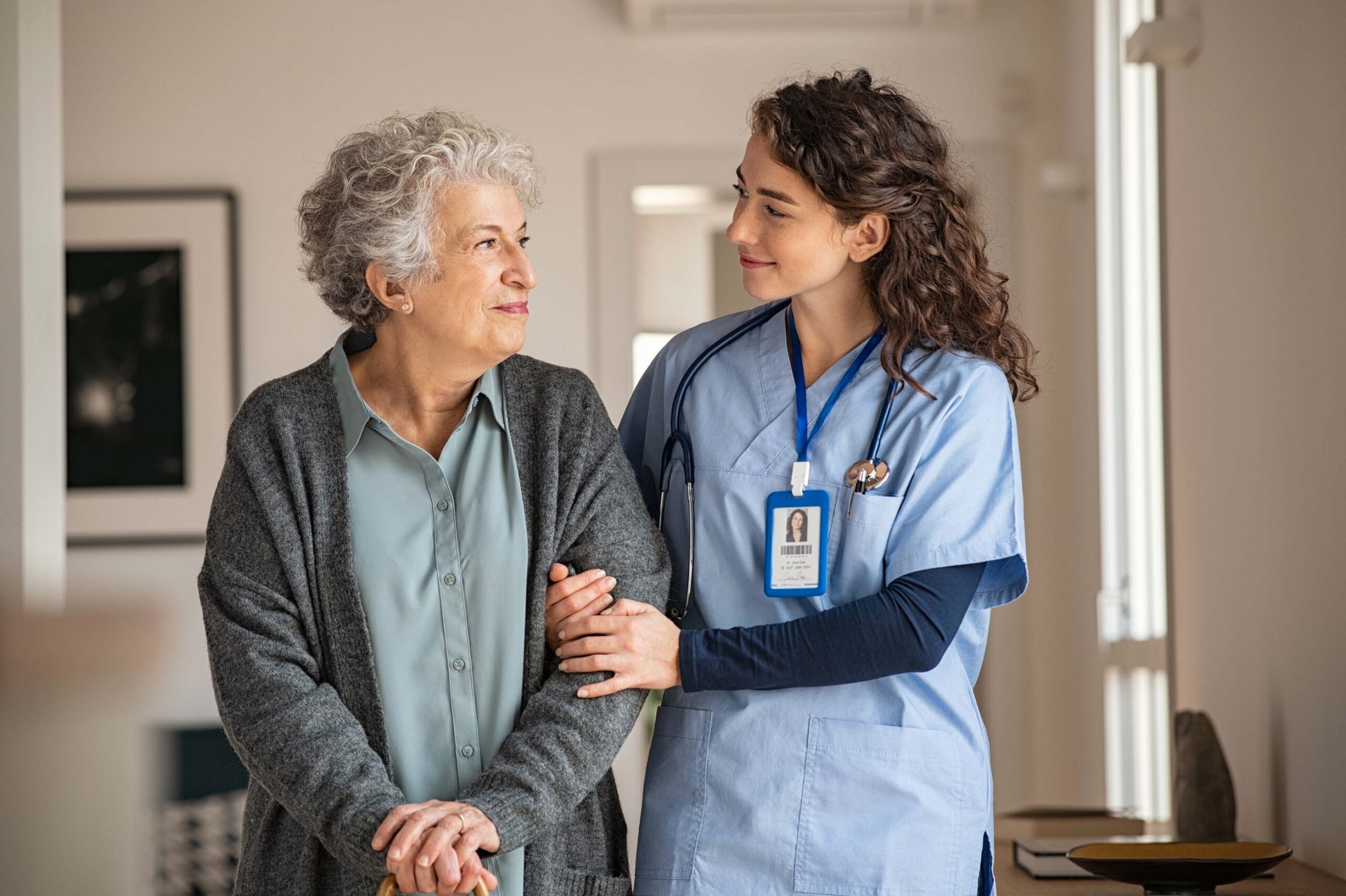 A nurse in blue scrubs with an ID badge gently supports an elderly woman with gray hair wearing a cardigan, representing the spirit of Care Initiative. They smile at each other warmly in a bright, home-like setting.