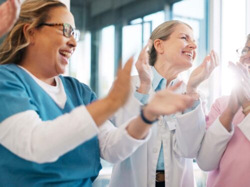 A group of healthcare professionals in scrubs and lab coats stands together indoors, smiling and clapping. Sunlight streams through large windows, creating a bright and positive atmosphere as they celebrate the successful launch of Care Initiative.