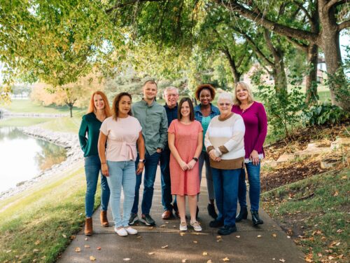 A group of eight people stands together on a tree-lined path by a pond, smiling in casual attire. Sunlight filters through the trees, creating a warm and inviting atmosphere. Their presence reflects a sense of unity and purpose, embodying the spirit of Care Initiative.