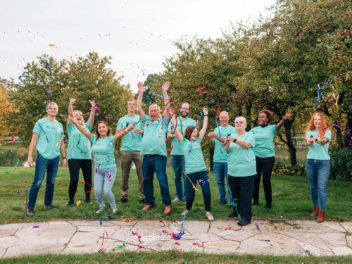 A group of people in light blue shirts celebrate outdoors on the grass, with trees in the background. As part of a Care Initiative, they smile and joyfully toss colorful streamers into the air.