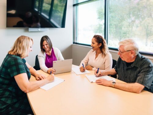 Four people are seated around a conference table in an office, engaged in a lively discussion about the new brand Care Initiative. Three are taking notes while one works on a laptop. A large screen is mounted on the wall behind them as natural light streams through the windows.