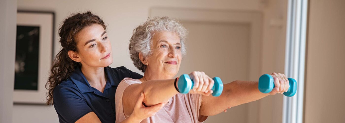 An older woman is doing arm exercises with blue dumbbells, assisted by a younger woman in a navy shirt. They are indoors near a window with natural light. The older woman is smiling and looks focused on her exercise, highlighting the Care Initiative they are engaged in.