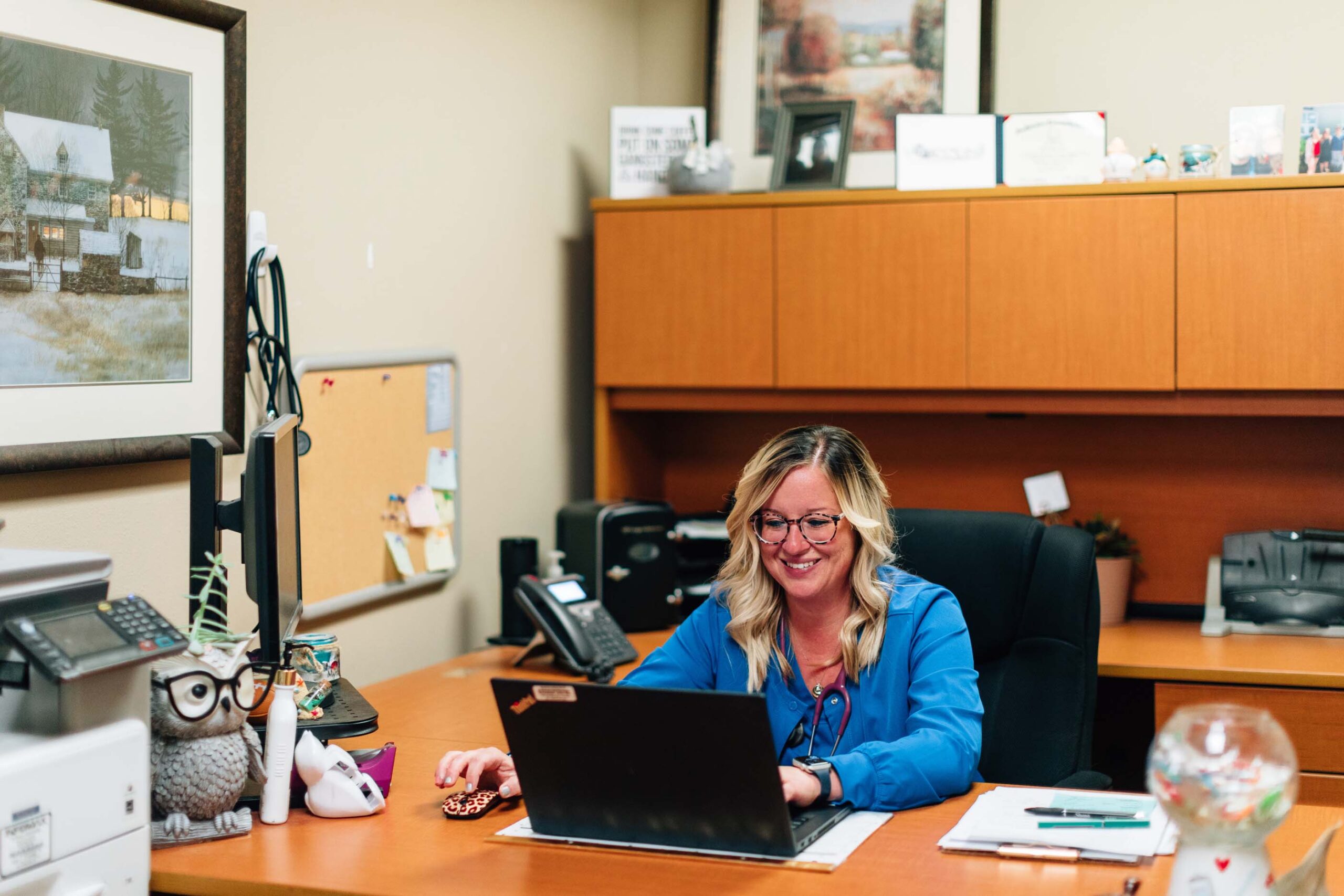 A woman in a blue blouse sits at a wooden desk, typing on a laptop. The desk features an owl figurine, a mouse, and a globe. Behind her are cabinets and framed artwork, symbolizing her focus on the Care Initiative in the workplace.