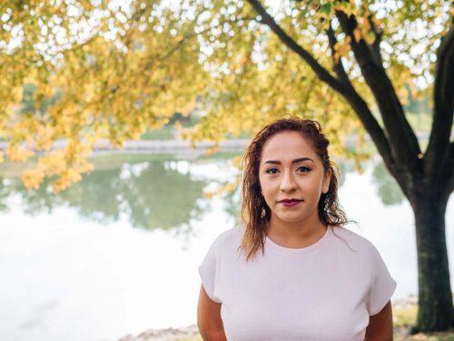 A woman with long hair stands by a lake, framed by autumn trees with golden leaves. She wears a white top and looks confidently at the camera. The Care Initiative enhances the serene backdrop as the water reflects the trees, creating a peaceful setting.