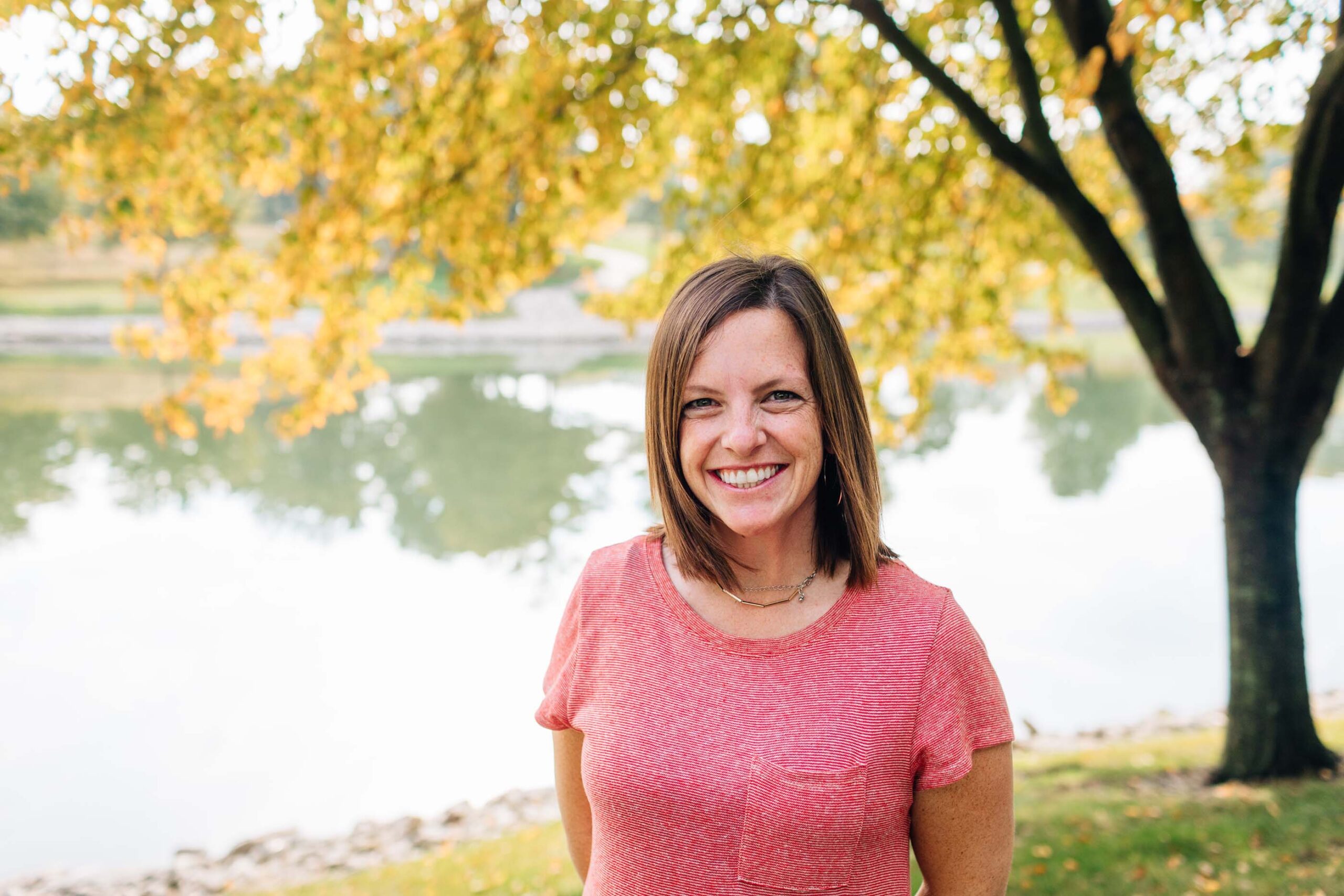 A woman in a pink shirt smiles by a serene lake with trees in vibrant autumn colors. A large tree with yellow leaves frames the scene, conveying calm and happiness on this pleasant day, embodying the essence of the Care Initiative by Care Initiative.