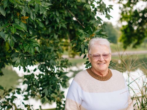 An older adult wearing glasses and a beige sweater smiles while standing outdoors. Green leaves and a blurred view of water create a peaceful, natural setting, embodying the essence of Care Initiative focused on well-being in nature.