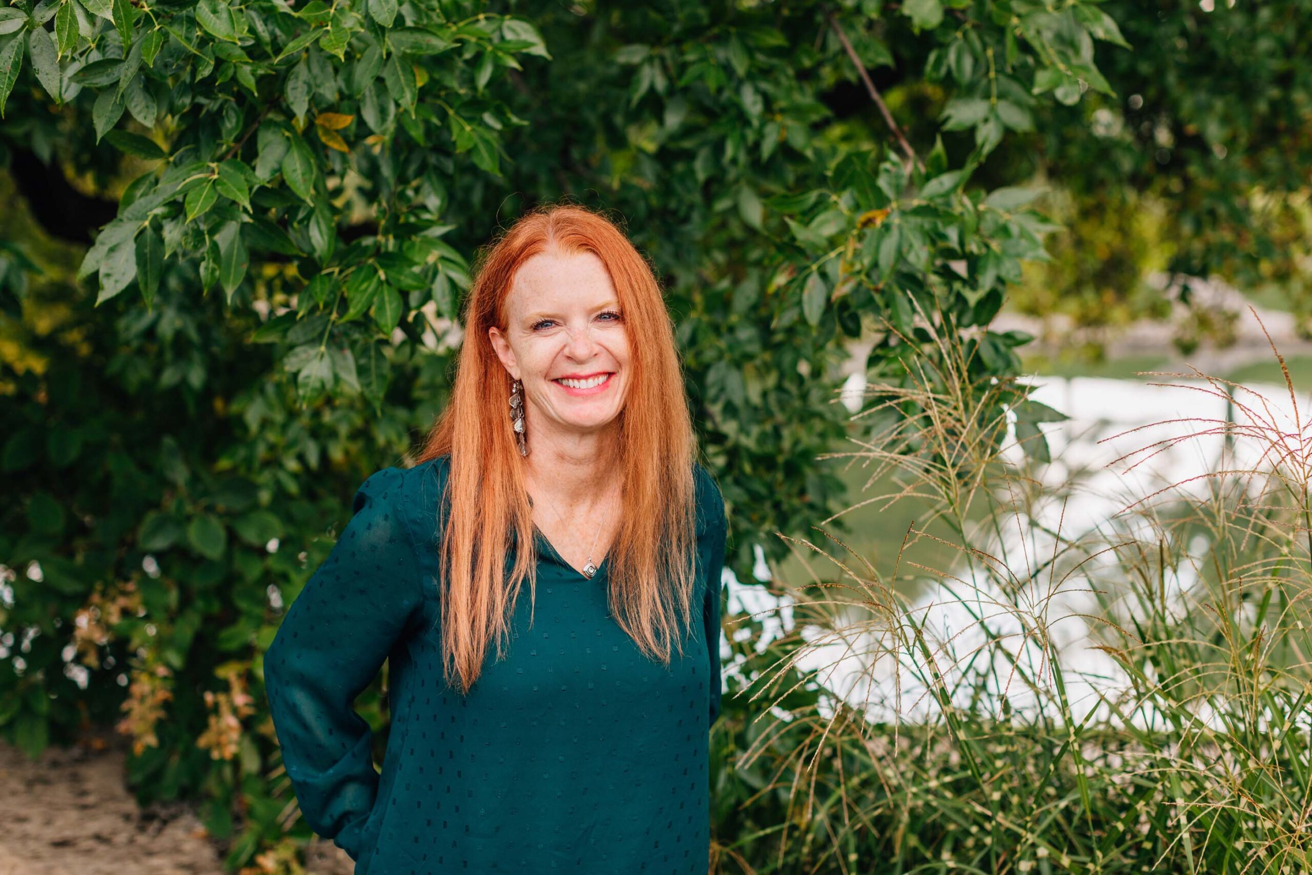 A woman with long red hair smiles while leaning against a tree. Wearing a green blouse, she stands amidst lush greenery with water visible in the background, embodying the spirit of Care Initiative for nature conservation.