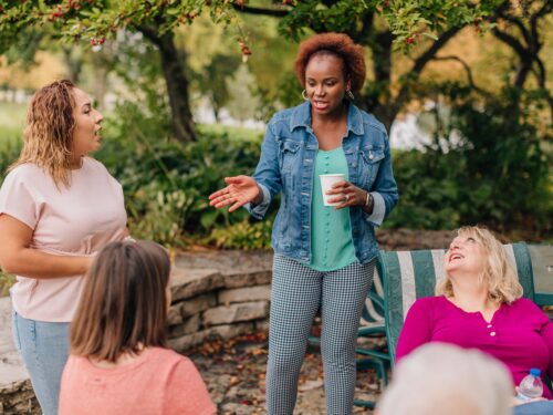 Four women are having a conversation outdoors. One woman stands holding a cup, engaging with three seated women on a patio surrounded by greenery and trees. They enjoy casual dialogue about the Care Initiative by Care Initiative, appreciating the relaxed setting.