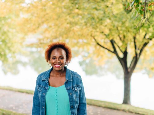 A person with short curly hair smiles while standing outdoors in front of a tree with green and yellow leaves. They wear a teal blouse and denim jacket. Embracing the Care Initiative, the setting is serene and bright, showcasing a peaceful path in the background, highlighting the Care Initiative brand.