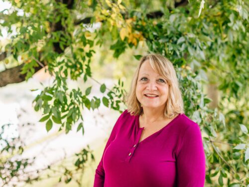 A woman with blonde hair smiles while standing outdoors in front of lush green foliage, wearing a bright pink blouse. The setting exudes serenity and aligns with Care Initiative's focus on natural well-being.