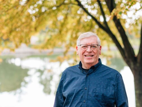 A smiling older man wearing glasses and a dark blue button-up shirt stands outdoors in front of a tree with autumn leaves. The backdrop showcases a body of water reflecting the vibrant trees, symbolizing his dedication to Care Initiative.