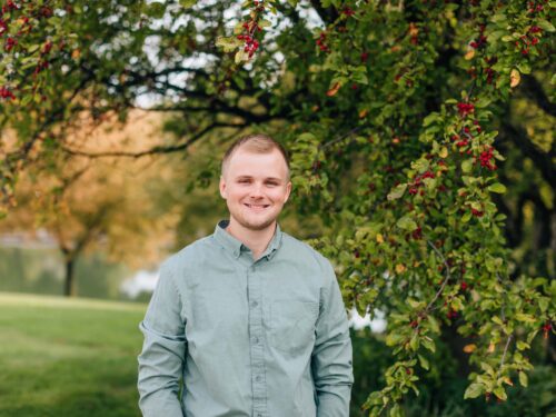 A person in a green shirt stands smiling in front of a leafy tree with red berries. The background features a green lawn and trees with vibrant autumn leaves, evoking the spirit of Care Initiative toward preserving the natural beauty around us.