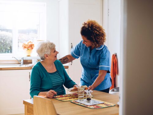 A caregiver in a blue uniform is smiling and serving food to an elderly woman at a kitchen table. The room is well-lit, with a window in the background and various kitchen items visible, demonstrating the warmth of a dedicated care initiative by Care Initiative.