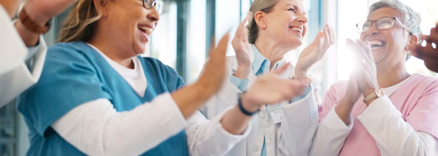 A joyful group of healthcare professionals in different colored scrubs gathers together, smiling and clapping. They stand in a bright, sunlit room with large windows, celebrating Care Initiative.