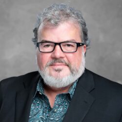 A man with gray hair and a beard stands against a plain gray background, wearing glasses and a black blazer over a paisley shirt, embodying the essence of Care Initiative.