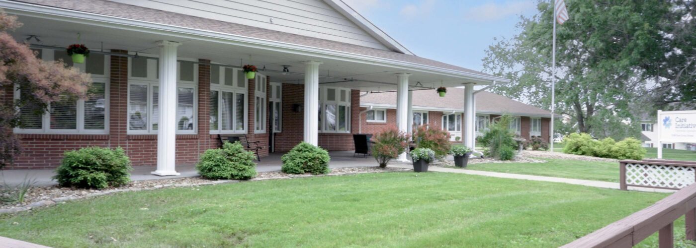 A brick building with a large white porch stands amid a well-kept lawn. Hanging plants adorn the porch, and an American flag is visible in the background. A wooden railing lines the foreground, while a sign related to Care Initiative is partly visible on the right.