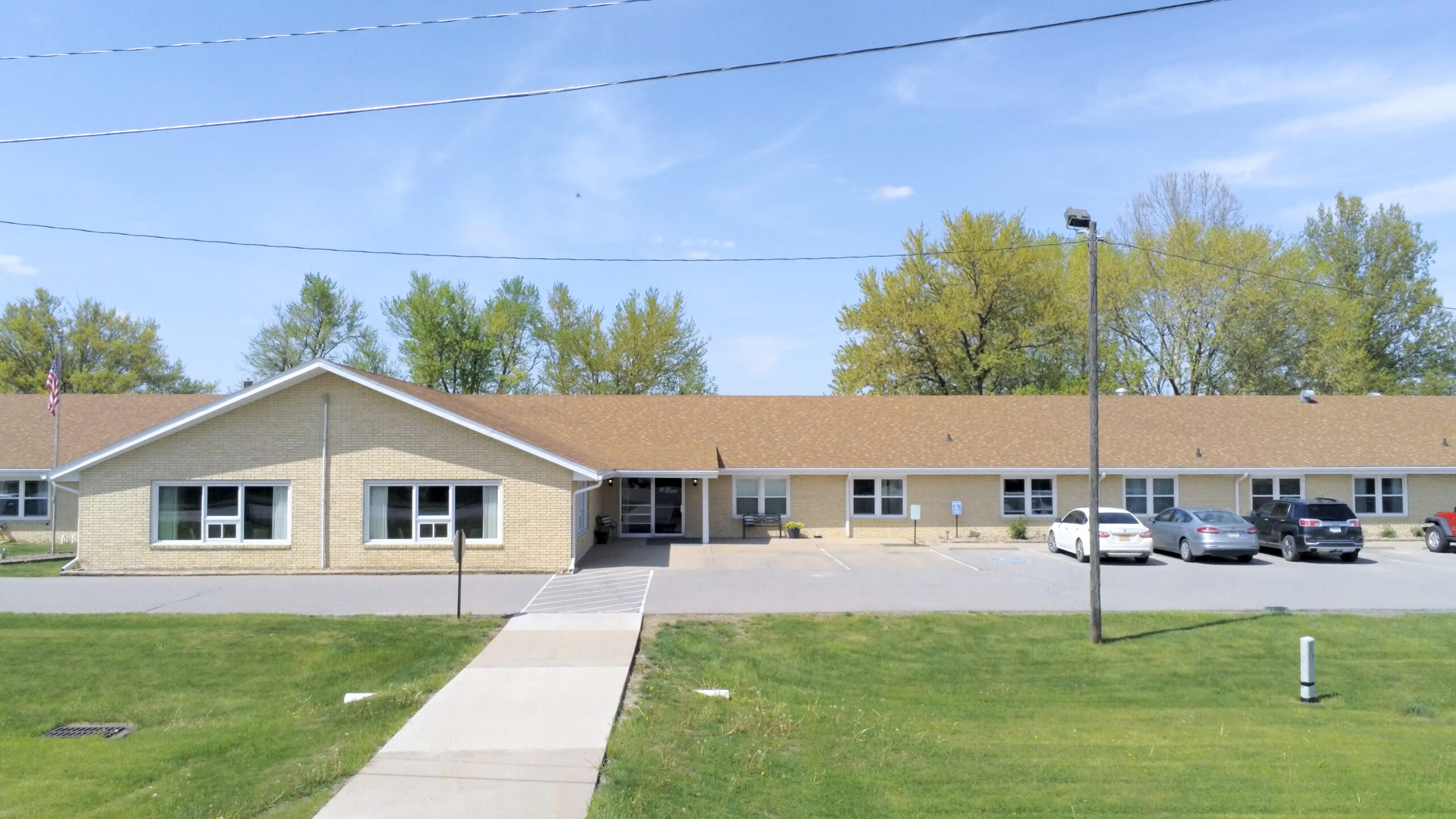 A single-story beige building with a brown roof and several windows stands amidst a paved parking lot with parked cars. A grassy lawn and concrete walkway lead to the entrance, reflecting its commitment to the Care Initiative. Trees can be seen in the background.