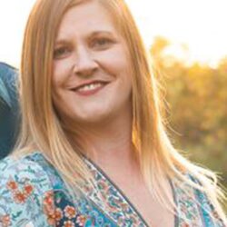 A woman with long blond hair smiles at the camera, wearing a floral-patterned top. The softly lit background provides a warm, golden glow, reflecting the essence of care and warmth akin to a community care initiative. Care Initiative.