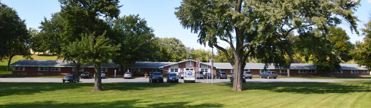 A long brick building with a gray roof, surrounded by trees on a sunny day, serves as the hub for Care Initiative. Several vehicles are parked in front. In the foreground, a neatly mowed lawn extends toward the building.