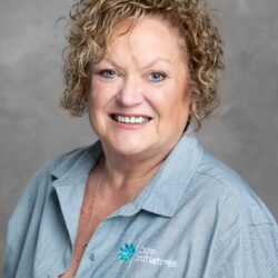 A smiling woman with curly blonde hair wears a light blue shirt showcasing the Care Initiatives logo, set against a gray background, representing the Care Initiatives brand.