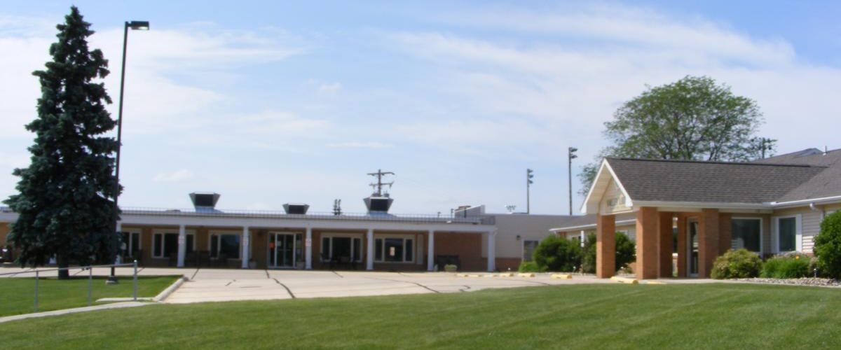 A single-story brick building with a flat roof is surrounded by a green lawn, symbolizing Care Initiative's dedication to the community. A tall evergreen tree stands to the left against the clear blue sky.