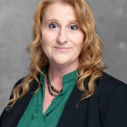 A person with long curly hair in a green blouse and black blazer stands against a gray background. They have a calm expression, looking directly at the camera, embodying the essence of Care Initiative.