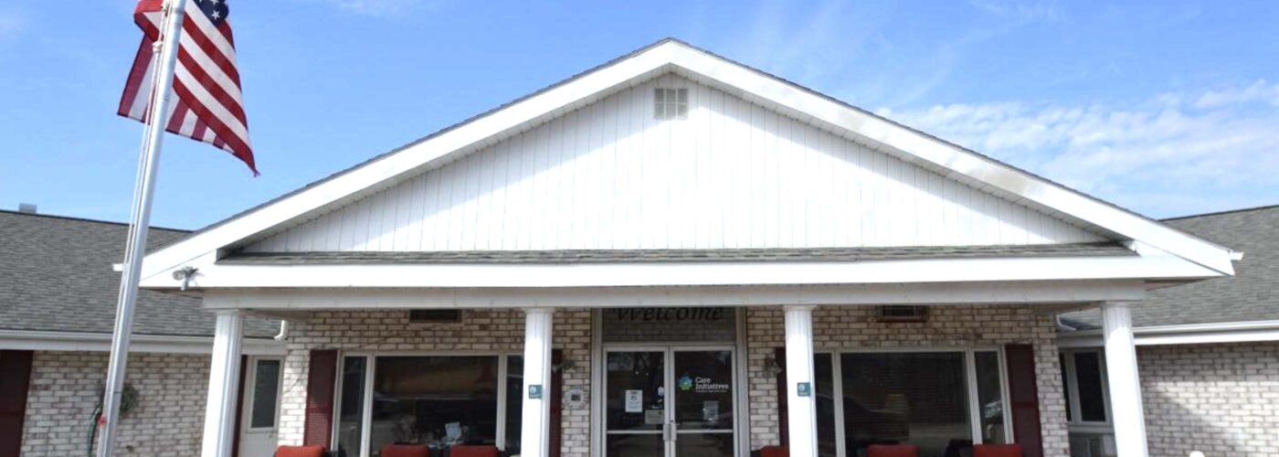 A single-story building with a gabled roof and a brick facade features a Welcome sign above the entrance. Red chairs sit on the porch, and an American flag is displayed on a flagpole under a bright blue sky, reflecting the spirit of Care Initiative.