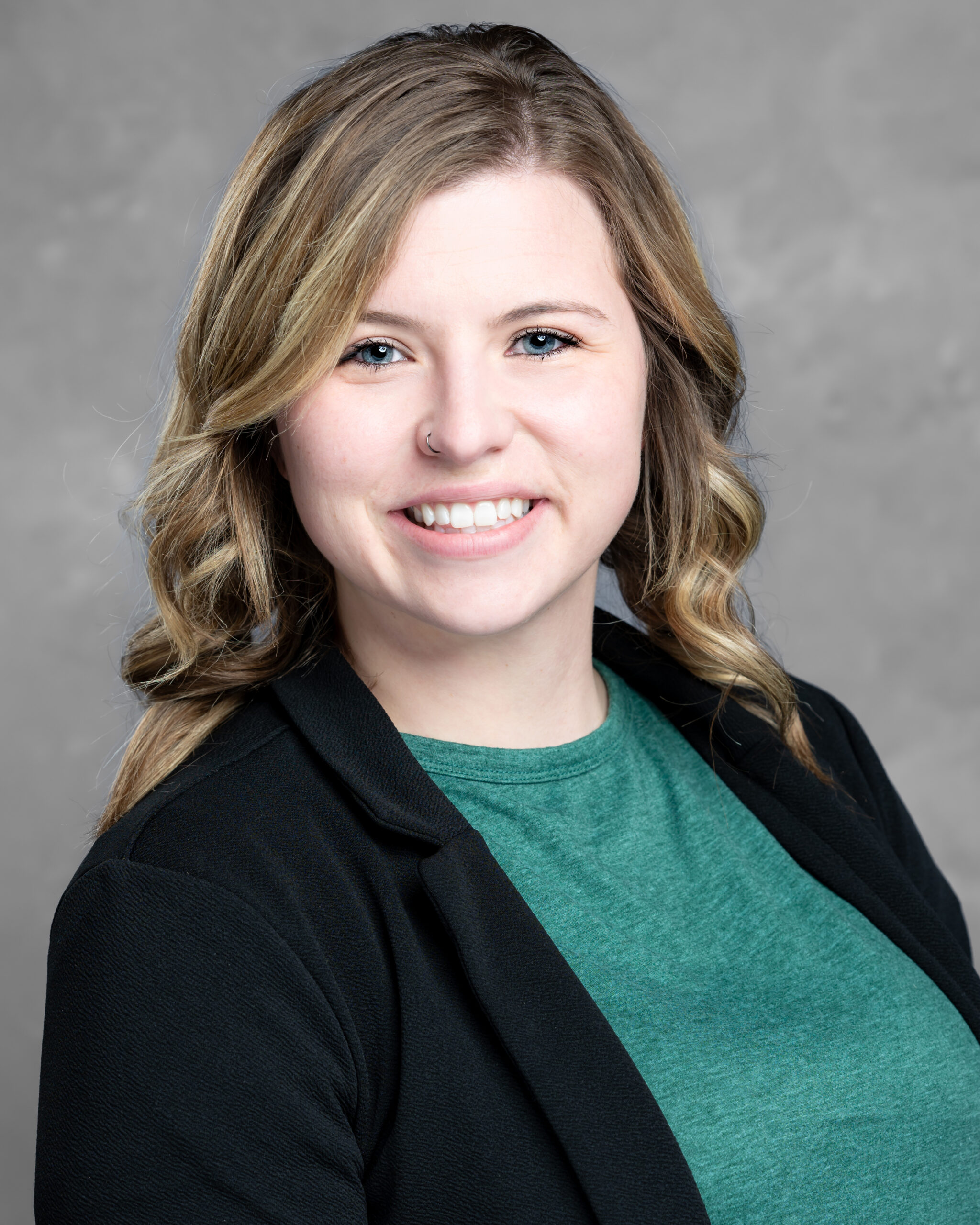 A smiling person with wavy blond hair wearing a green top and black blazer stands against a gray background, embodying the spirit of Care Initiative.