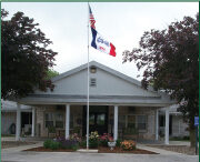 A single-story building with a flagpole displaying the American and Iowa state flags. The entrance is adorned with columns and surrounded by landscaped greenery and flowers, embodying a thoughtful care initiative by Care Initiative.