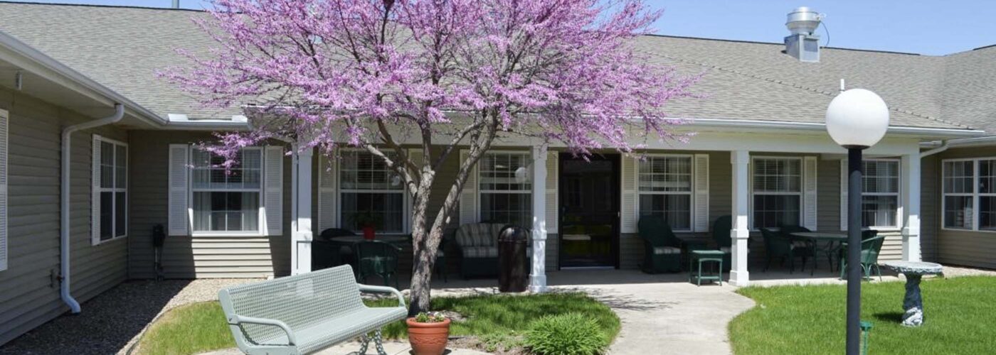 A courtyard with a blooming pink tree at its center hosts a green bench and potted plants along a pathway leading to a building with a shaded porch. The well-tended grass reflects the Care Initiative, and a lamppost graces the entrance nearby.