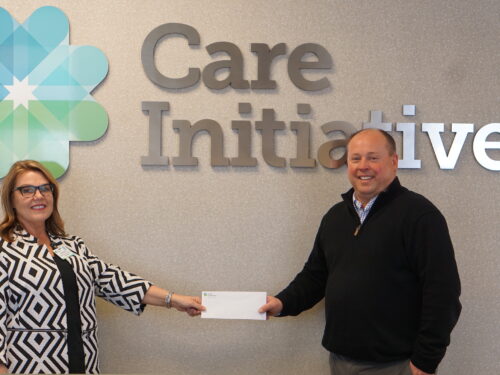 Two individuals stand before a wall displaying the Care Initiatives logo, smiling as they hold an envelope. The person on the left is dressed in a patterned outfit, while the one on the right wears a dark sweater. The brand name Care Initiative is prominently featured behind them.