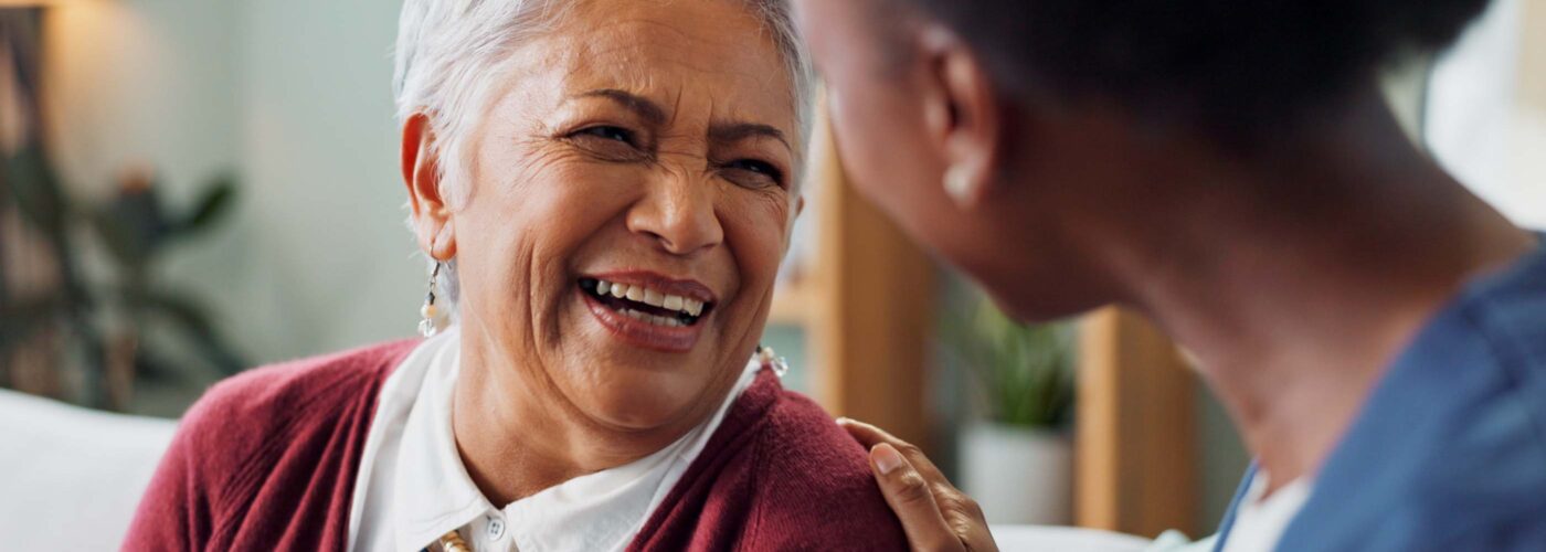 An elderly woman with short gray hair and a red cardigan laughs while sitting on a couch. Beside her, a younger person is smiling and touches her shoulder in support, embodying the spirit of Care Initiative that brings generations together.